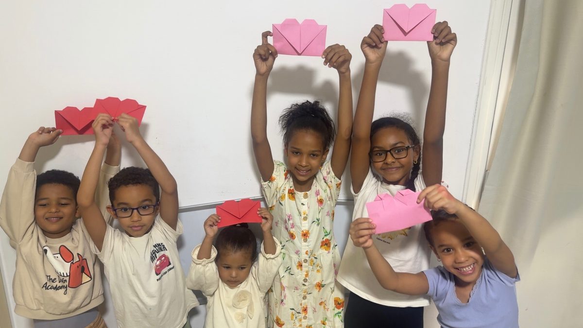 Seven children in a classroom with whiteboards hold red and pink heart envelopes over their heads, smiling.