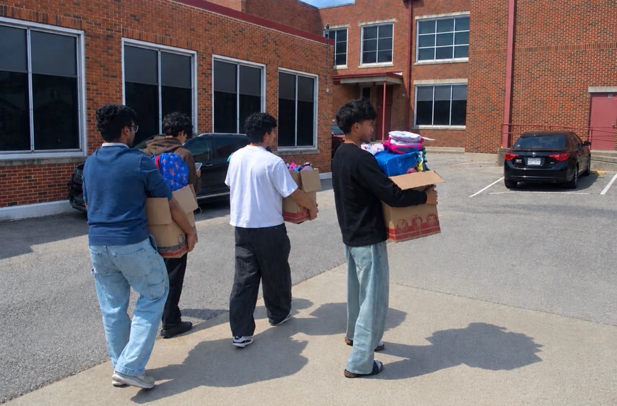 Four team members carry cardboard boxes of supplies across a parking lot toward a brick school building.