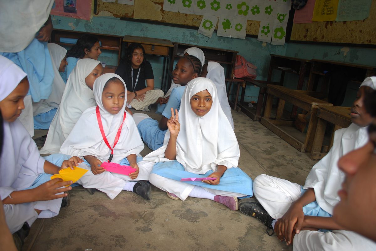 A classroom of girls in white headscarves seated in a circle, opening heart envelopes.