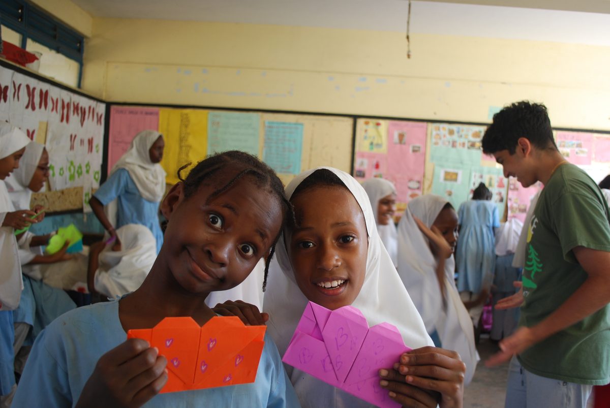 Two girls stand shoulder to shoulder holding envelopes covered in hand-drawn hearts, one tilting her head with wide eyes, the other beaming at the camera.