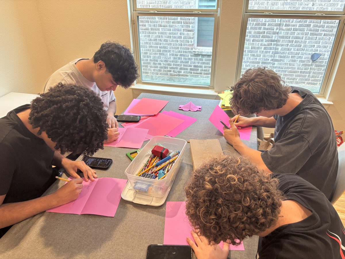 Overhead view of four teen volunteers writing letters on pink paper, a folded purple heart envelope at the center of the table.