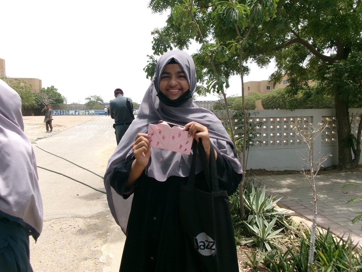A girl in a grey hijab laughs while holding a pink envelope decorated with hand-drawn hearts, a campus wall and trees behind her.